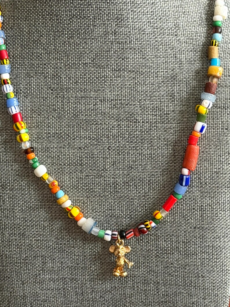 May include: A colorful beaded necklace with a gold-colored Mickey Mouse charm. The necklace features a variety of small, multi-colored beads, including red, yellow, blue, and white. The charm is a detailed figure of Mickey Mouse.