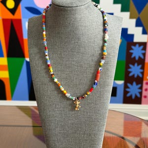 May include: A colorful beaded necklace with a gold-colored charm of a mouse. The necklace features a variety of small, multi-colored beads, including red, blue, yellow, and white. The necklace is displayed on a gray mannequin.