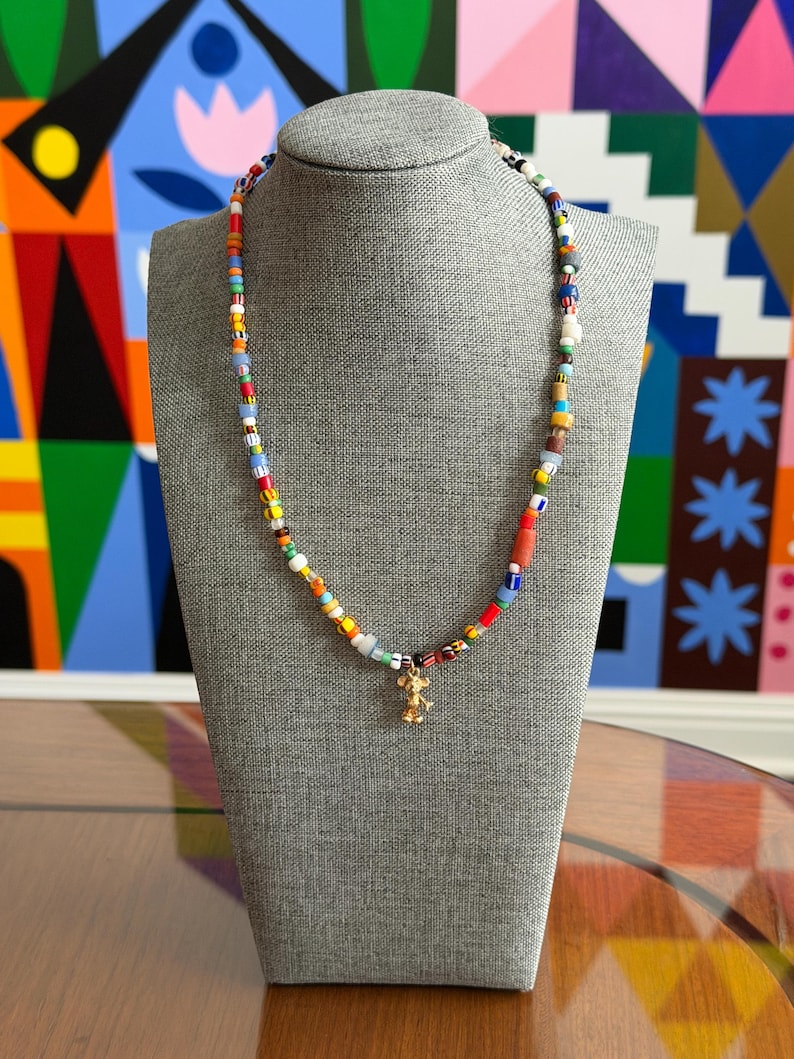 May include: A colorful beaded necklace with a gold-colored charm of a mouse. The necklace features a variety of small, multi-colored beads, including red, blue, yellow, and white. The necklace is displayed on a gray mannequin.