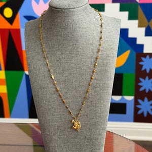 May include: A gold-colored necklace with a delicate chain and a detailed pendant. The necklace is displayed on a gray mannequin against a colorful abstract background. The pendant appears to be a floral or ornate design.