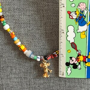 May include: A colorful beaded necklace with a gold-colored Mickey Mouse charm. The necklace features a variety of small, round beads in multiple colors. A ruler is placed next to the necklace for scale.