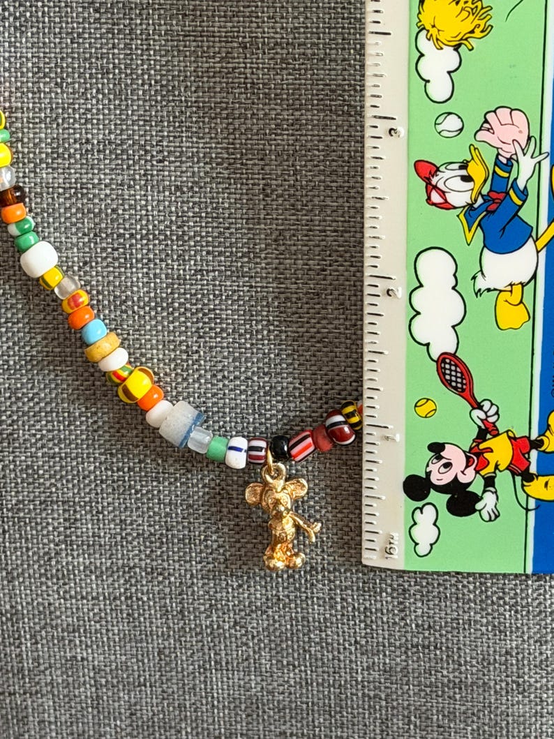 May include: A colorful beaded necklace with a gold-colored Mickey Mouse charm. The necklace features a variety of small, round beads in multiple colors. A ruler is placed next to the necklace for scale.