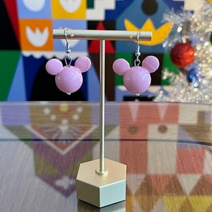Puede incluir: Un par de pendientes de cabeza de Mickey Mouse de color rosa con ganchos plateados que cuelgan de un soporte dorado.