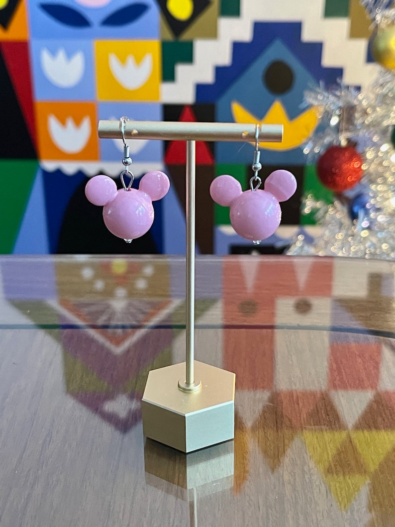 Puede incluir: Un par de pendientes de cabeza de Mickey Mouse de color rosa con ganchos plateados que cuelgan de un soporte dorado.