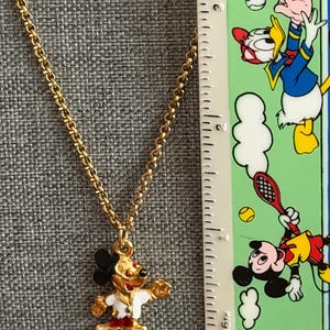 K&ouml;nnte beinhalten: Goldfarbene Halskette mit einem Mickey Mouse Anh&auml;nger. Der Anh&auml;nger ist rot, wei&szlig;, schwarz und goldfarben emailliert. Die Kette ist im filigranen Glieder-Stil gehalten. Ein Lineal mit Cartoon-Figuren ist im Hintergrund zu sehen.