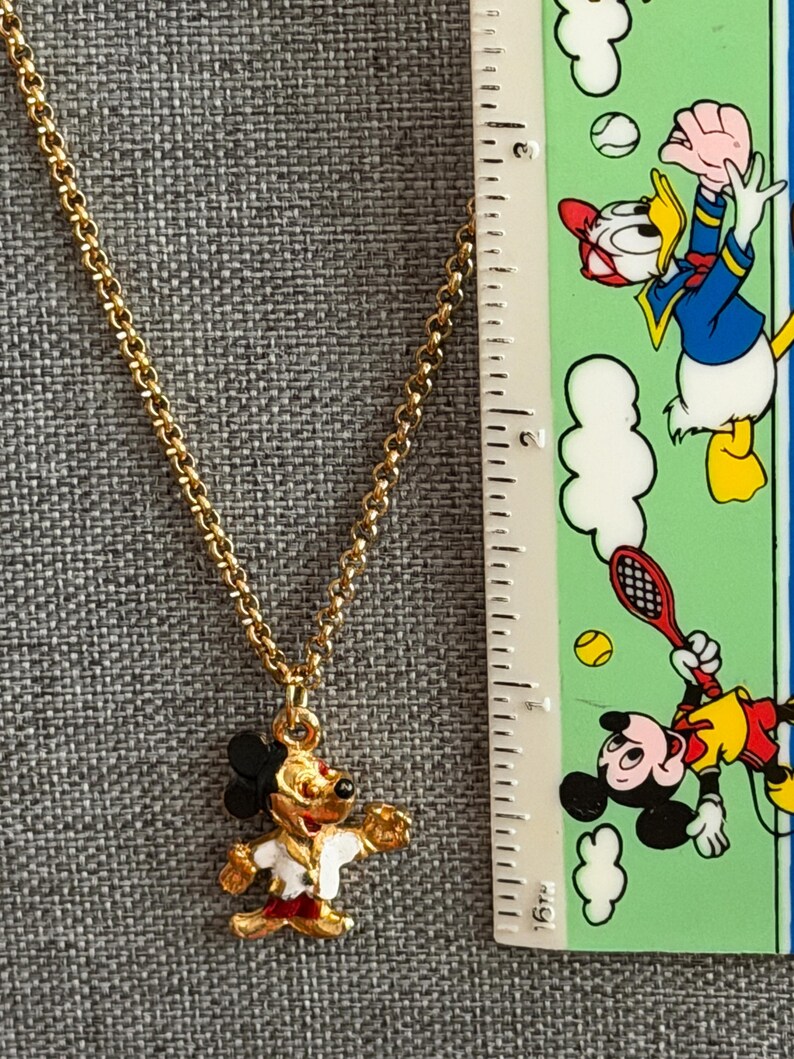 K&ouml;nnte beinhalten: Goldfarbene Halskette mit einem Mickey Mouse Anh&auml;nger. Der Anh&auml;nger ist rot, wei&szlig;, schwarz und goldfarben emailliert. Die Kette ist im filigranen Glieder-Stil gehalten. Ein Lineal mit Cartoon-Figuren ist im Hintergrund zu sehen.