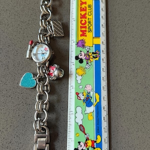 Puede incluir: Reloj de pulsera plateado con esfera redonda y varios abalorios, incluyendo un labial, un coraz&oacute;n y un bolso. La esfera del reloj presenta un dise&ntilde;o de dibujos animados. Una regla de Mickey Sport Club est&aacute; al lado del reloj.