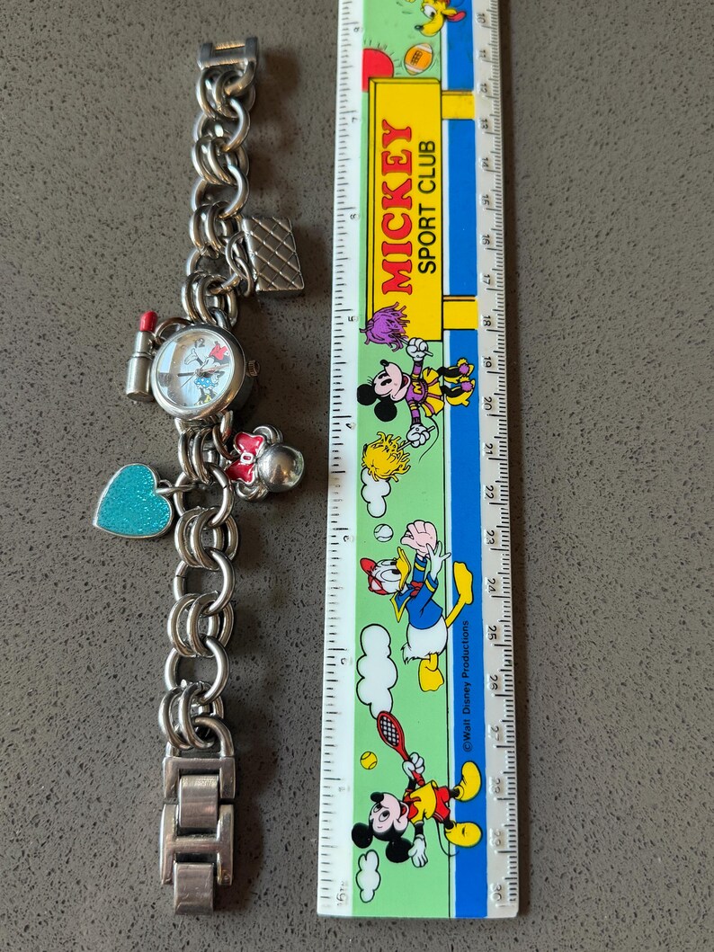 Puede incluir: Reloj de pulsera plateado con esfera redonda y varios abalorios, incluyendo un labial, un coraz&oacute;n y un bolso. La esfera del reloj presenta un dise&ntilde;o de dibujos animados. Una regla de Mickey Sport Club est&aacute; al lado del reloj.