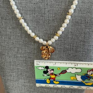 May include: A pearl necklace with gold accents and a gold Mickey Mouse pendant. The necklace is displayed on a gray surface. A ruler with a Mickey Mouse graphic is in the image.