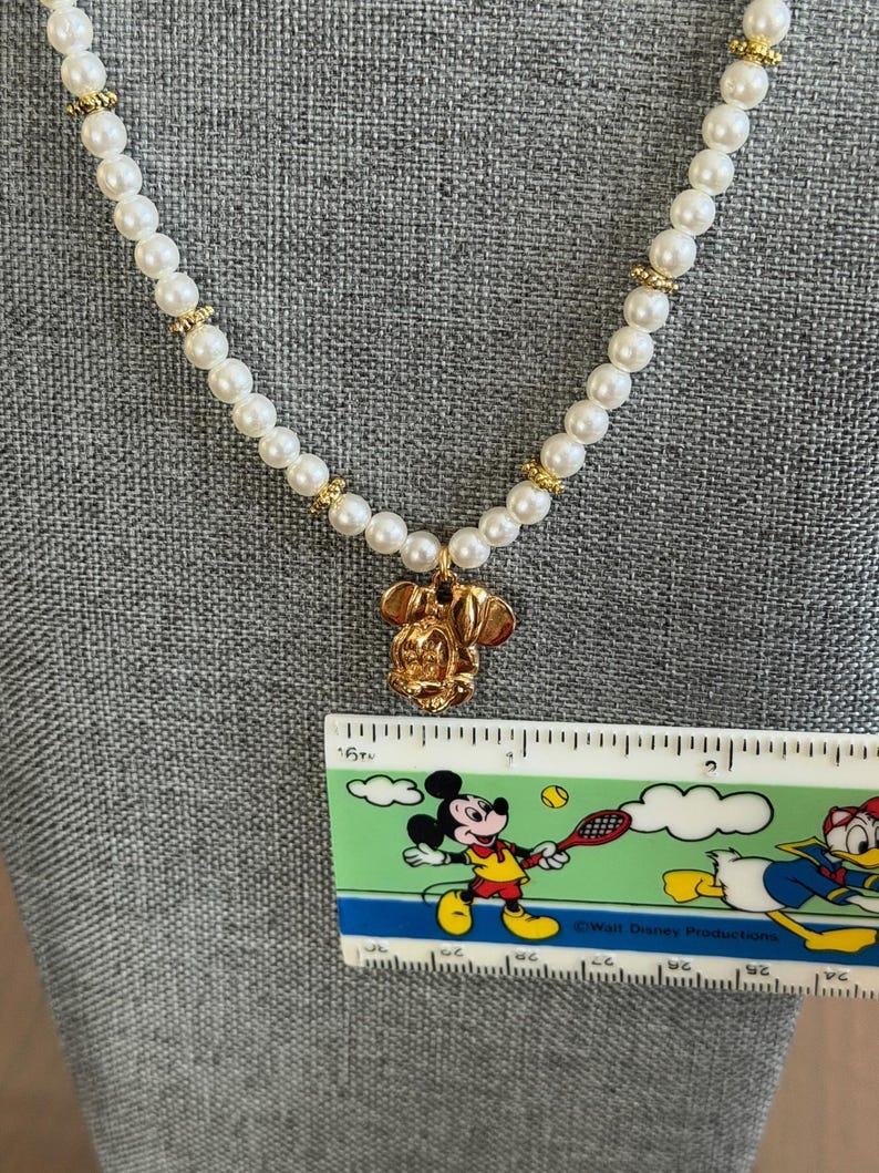 May include: A pearl necklace with gold accents and a gold Mickey Mouse pendant. The necklace is displayed on a gray surface. A ruler with a Mickey Mouse graphic is in the image.