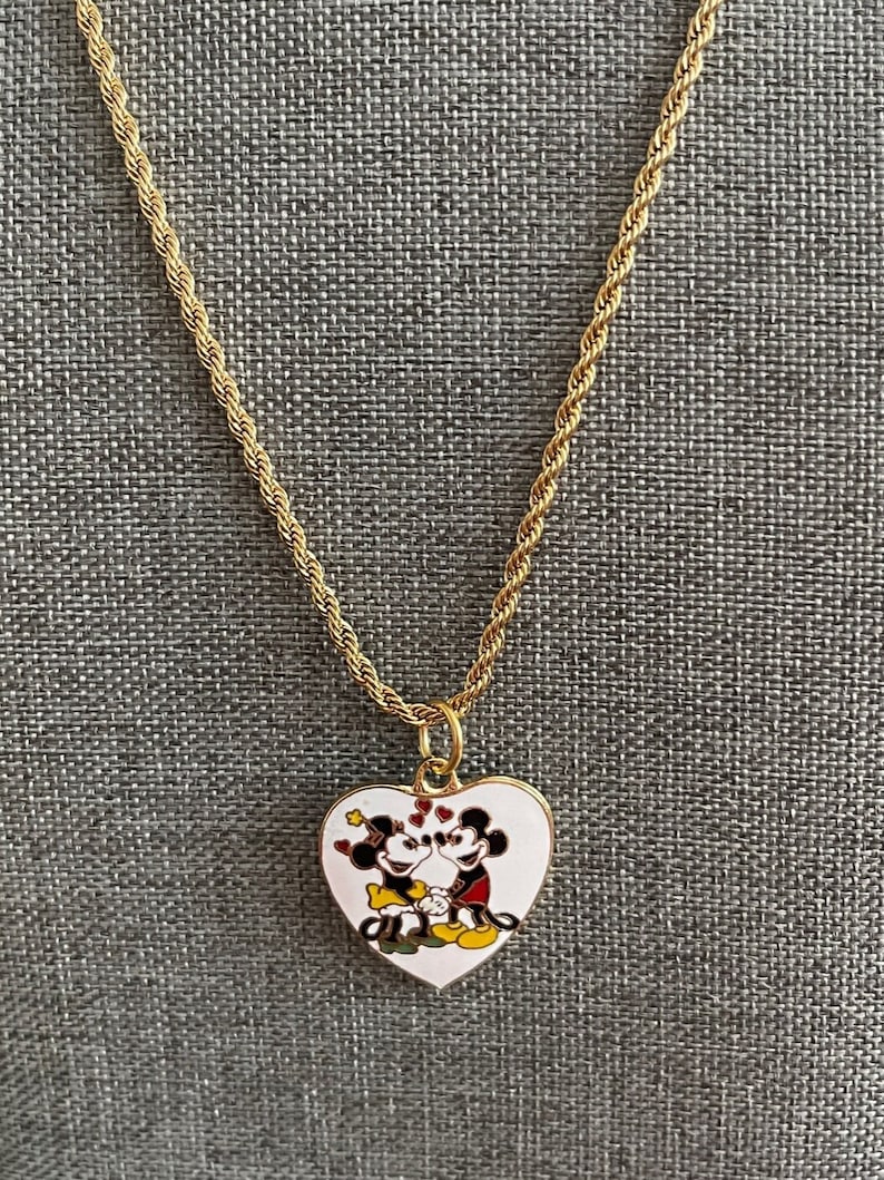 May include: A gold chain necklace with a heart-shaped pendant featuring a cartoon image of Mickey Mouse and Minnie Mouse kissing. The pendant is white with red, black, and yellow accents.