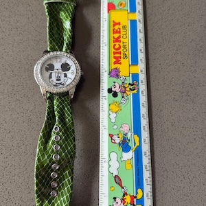 May include: A silver-toned wristwatch with a white face featuring Mickey Mouse. The watch has a bezel encrusted with rhinestones and a green, snakeskin-patterned band. The watch is next to a ruler with Mickey Mouse characters.