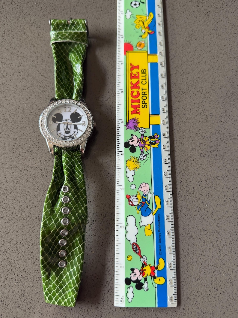 May include: A silver-toned wristwatch with a white face featuring Mickey Mouse. The watch has a bezel encrusted with rhinestones and a green, snakeskin-patterned band. The watch is next to a ruler with Mickey Mouse characters.