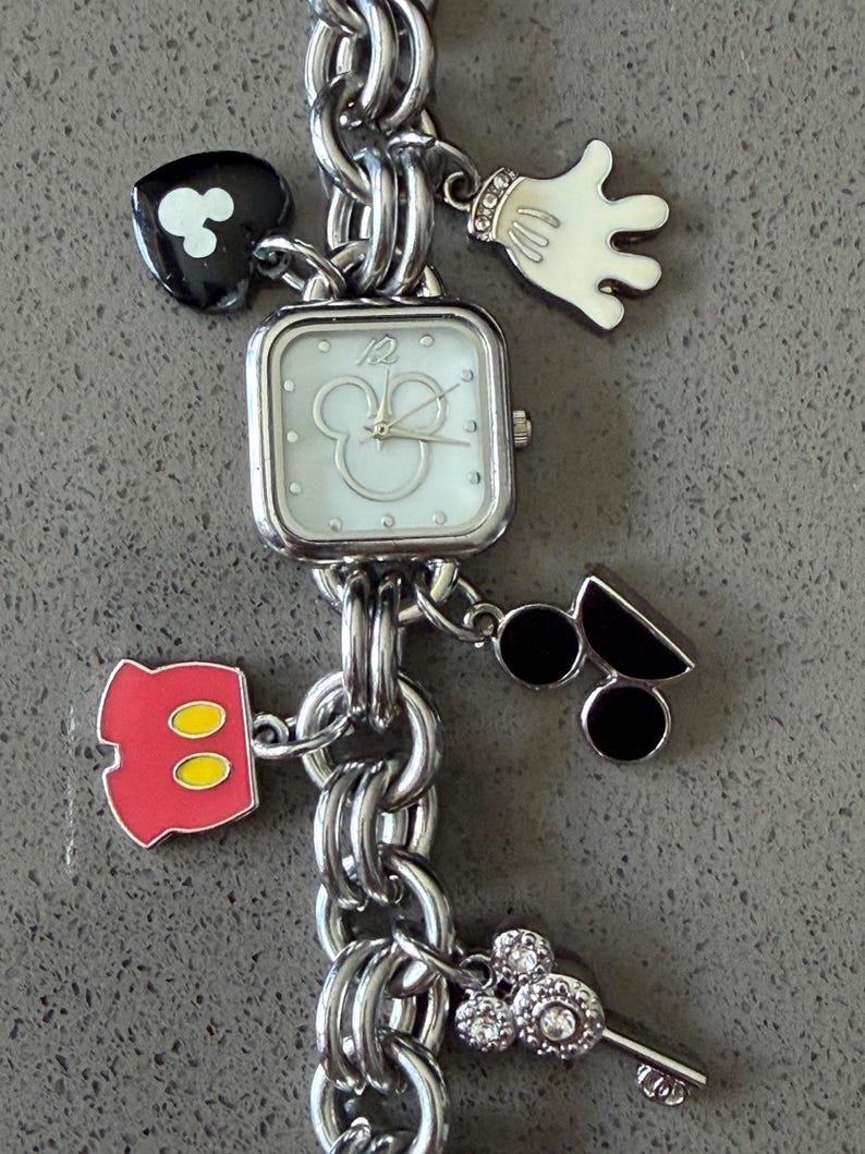 May include: Silver-toned charm bracelet watch featuring Mickey Mouse-themed charms. The watch has a square face with a Mickey Mouse silhouette. Charms include a Mickey glove, shorts, and a key. The bracelet is made of linked metal.