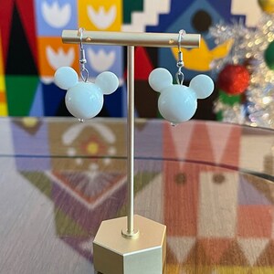 Puede incluir: Pendientes blancos con forma de cabeza de Mickey Mouse en un soporte dorado. Los pendientes est&aacute;n en un gancho plateado.
