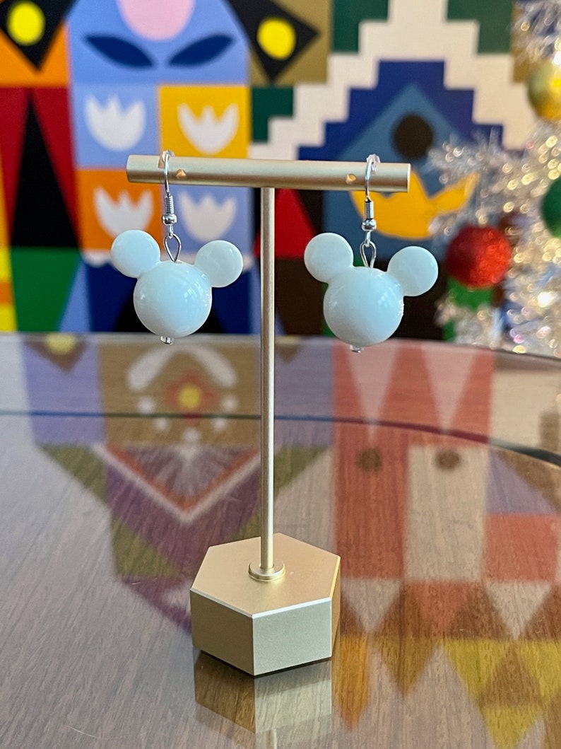 Puede incluir: Pendientes blancos con forma de cabeza de Mickey Mouse en un soporte dorado. Los pendientes est&aacute;n en un gancho plateado.