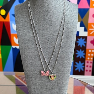May include: Two silver necklaces displayed on a gray mannequin. One necklace features a pink bow pendant with white polka dots. The other necklace has a heart-shaped pendant with a Minnie Mouse design.