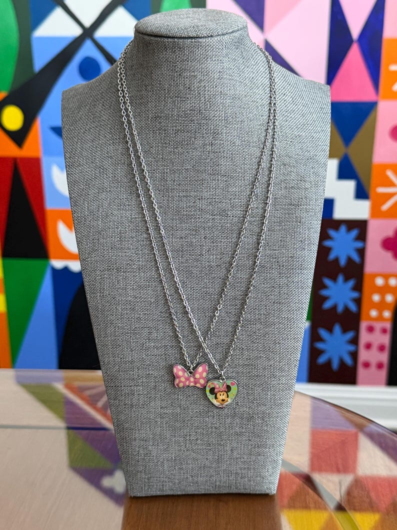 May include: Two silver necklaces displayed on a gray mannequin. One necklace features a pink bow pendant with white polka dots. The other necklace has a heart-shaped pendant with a Minnie Mouse design.