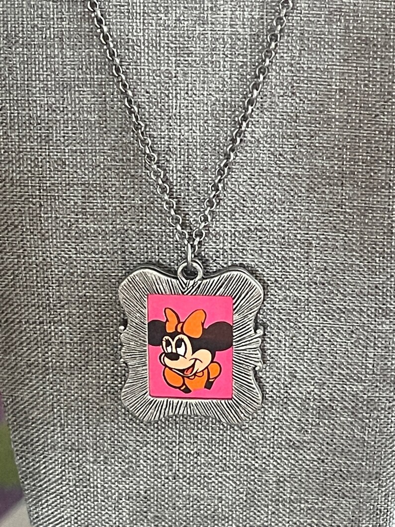 May include: A silver chain necklace with a silver frame pendant featuring a cartoon image of Minnie Mouse with a pink background and orange bow.