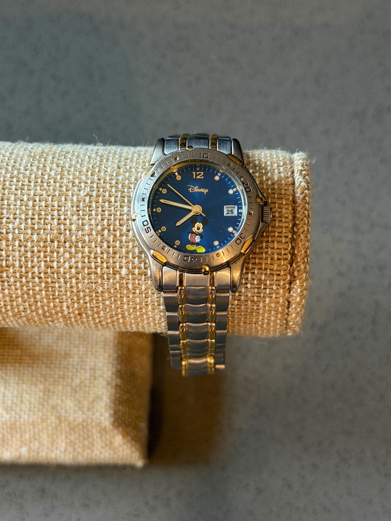 Op de afbeelding: Een zilver- en goudkleurig Disney-horloge met een blauwe wijzerplaat met Mickey Mouse. Het horloge heeft een datumweergave op 3 uur en een roestvrijstalen band. Het horloge wordt getoond op een beige stof.