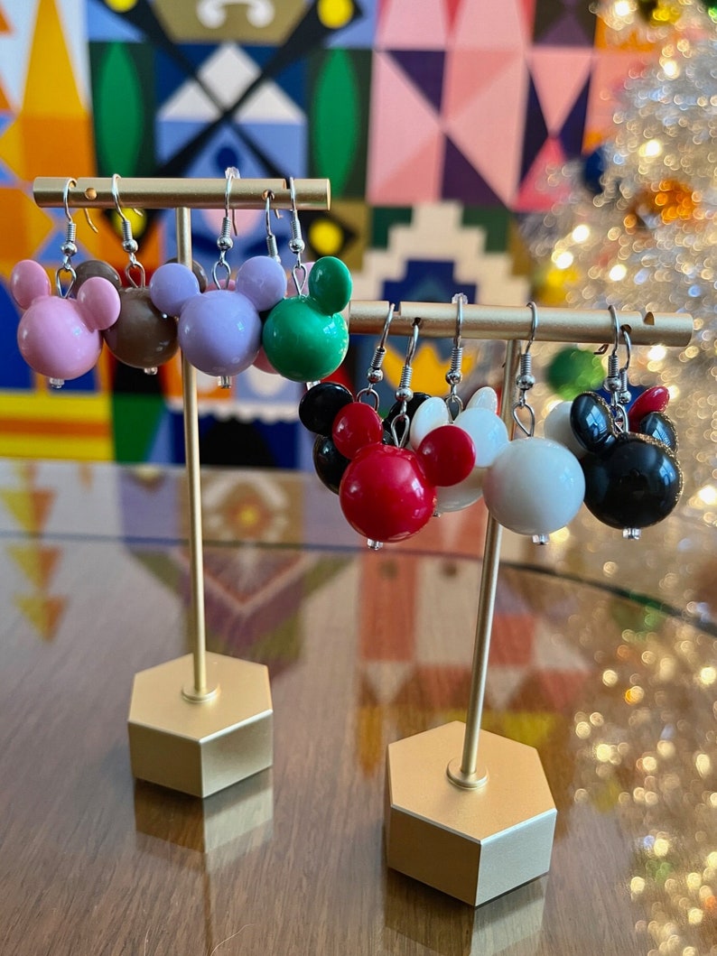 Puede incluir: Seis pares de pendientes colgantes con un dise&ntilde;o de cabeza de Mickey Mouse. Los pendientes est&aacute;n hechos de pl&aacute;stico y vienen en una variedad de colores, incluyendo rosa, marr&oacute;n, morado, verde, rojo, blanco y negro. Los pendientes se muestran en un soporte dorado.