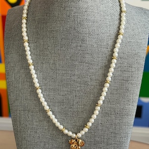 May include: A pearl necklace with gold accents and a gold pendant in the shape of a cartoon character's head. The necklace is displayed on a gray mannequin.