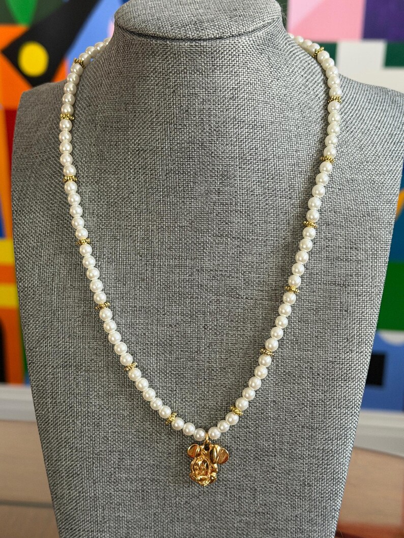 May include: A pearl necklace with gold accents and a gold pendant in the shape of a cartoon character's head. The necklace is displayed on a gray mannequin.