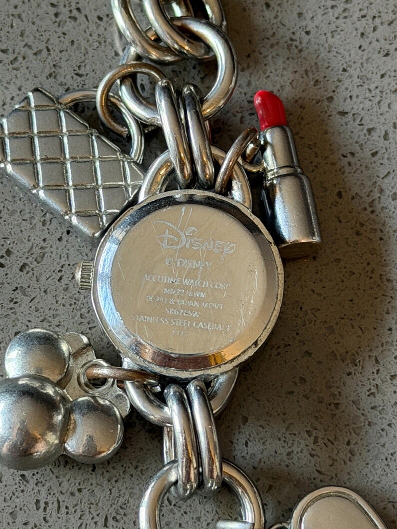Puede incluir: Pulsera de dijes plateada con una esfera de reloj de Disney y varios dijes. Los dijes incluyen un l&aacute;piz labial, una cabeza de Mickey Mouse y un dise&ntilde;o en forma de diamante. La esfera del reloj tiene el logotipo de Disney y texto.