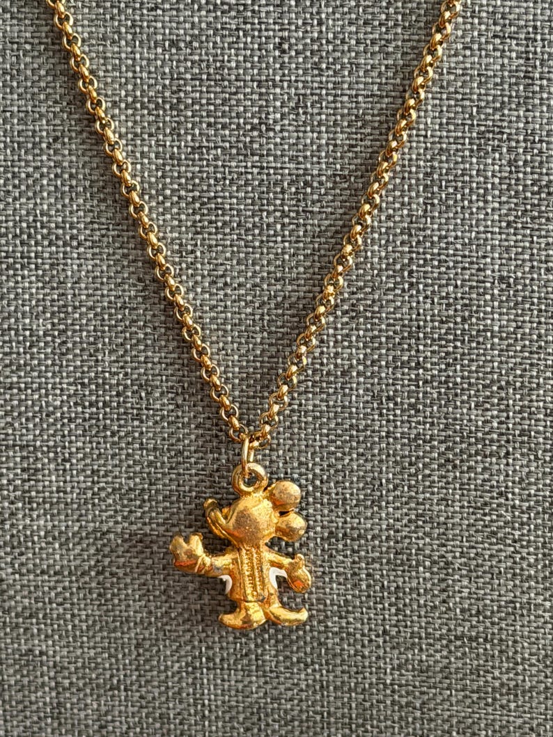 K&ouml;nnte beinhalten: Eine goldfarbene Halskette mit einem Anh&auml;nger in Form einer Zeichentrickmaus. Der Anh&auml;nger hat ausgestreckte Arme und ein l&auml;chelndes Gesicht. Die Kette ist zart goldfarben.