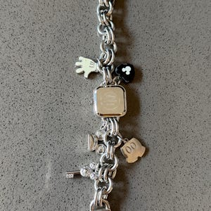 May include: Silver-toned charm bracelet with a square watch face. The bracelet features various charms, including a white glove, a black flower, a film camera, a key, and a Mickey Mouse silhouette. The watch face has the Disney logo.