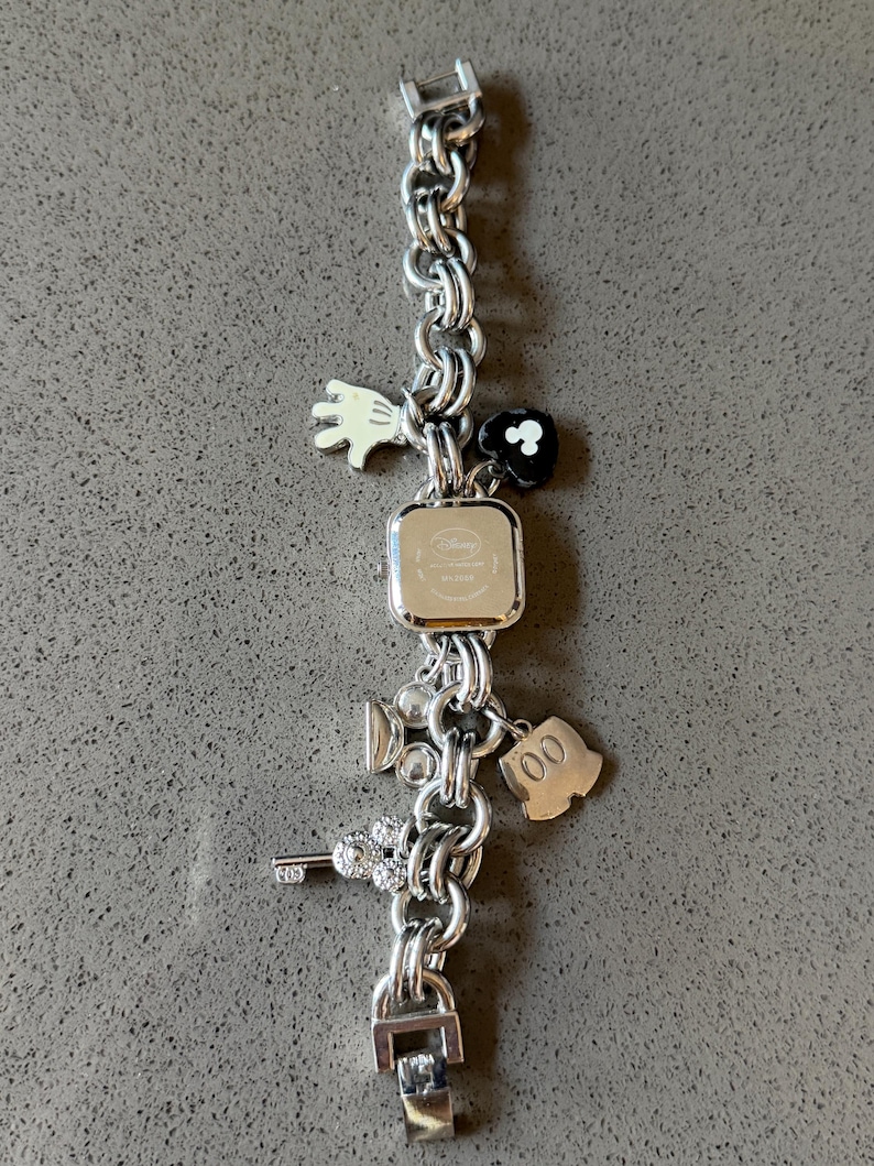 May include: Silver-toned charm bracelet with a square watch face. The bracelet features various charms, including a white glove, a black flower, a film camera, a key, and a Mickey Mouse silhouette. The watch face has the Disney logo.