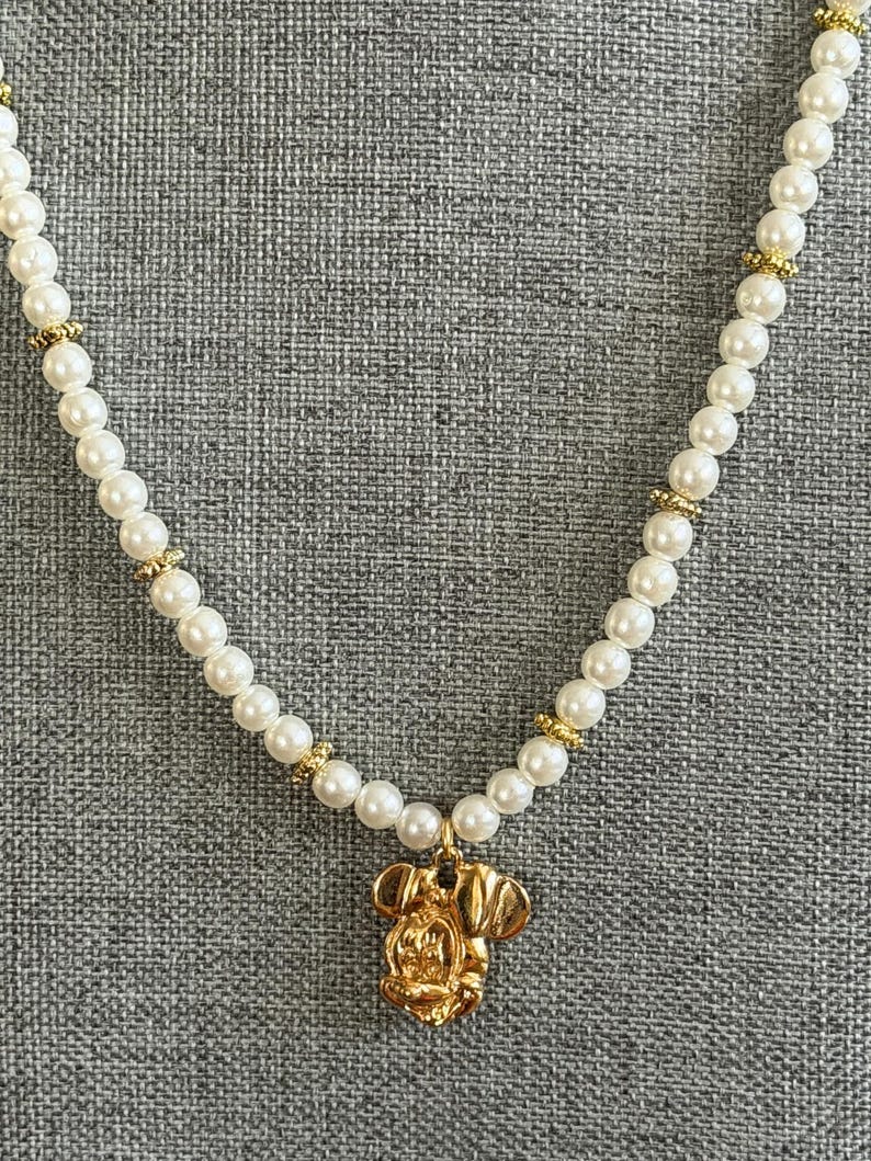 May include: A pearl necklace with a gold pendant in the shape of a cartoon character's head. The necklace features white, round pearls and gold-colored accents. The pendant is a detailed, gold-colored charm.