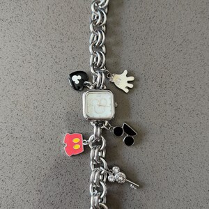 May include: Silver-toned charm bracelet watch with a square face and a chain-link band. The watch features Mickey Mouse-themed charms, including a heart, glove, and silhouette of Mickey's head. The watch is approximately 7 inches long.