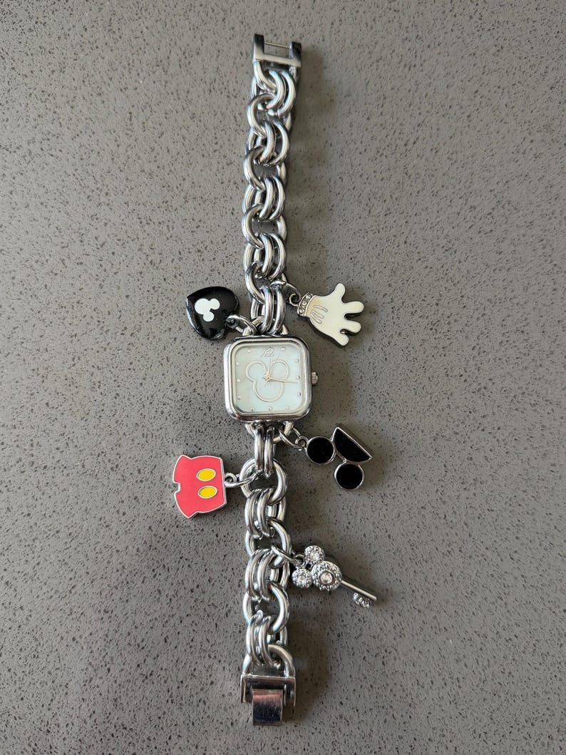 May include: Silver-toned charm bracelet watch with a square face and a chain-link band. The watch features Mickey Mouse-themed charms, including a heart, glove, and silhouette of Mickey's head. The watch is approximately 7 inches long.