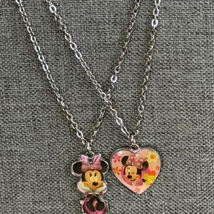 Peut inclure: Deux colliers argent&eacute;s avec des pendentifs Minnie Mouse. L'un est une figurine de Minnie Mouse, l'autre un c&oelig;ur rose avec un motif Minnie Mouse et des d&eacute;tails floraux. Les colliers sont pr&eacute;sent&eacute;s sur un tissu gris.