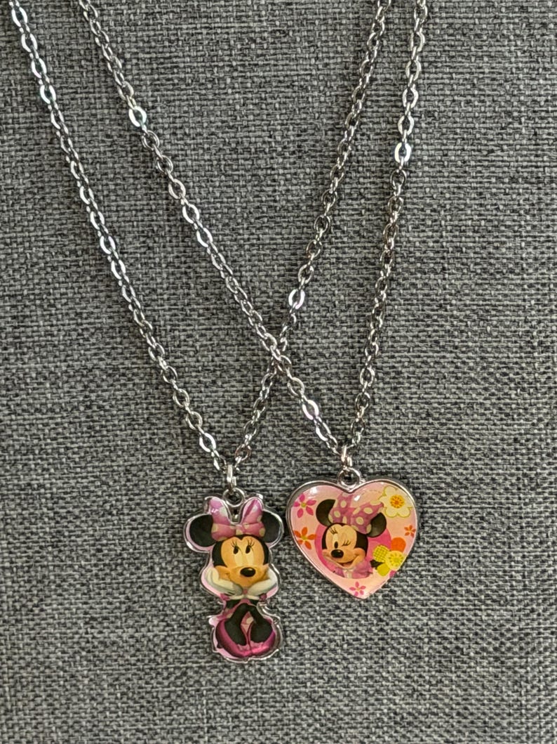Peut inclure: Deux colliers argent&eacute;s avec des pendentifs Minnie Mouse. L'un est une figurine de Minnie Mouse, l'autre un c&oelig;ur rose avec un motif Minnie Mouse et des d&eacute;tails floraux. Les colliers sont pr&eacute;sent&eacute;s sur un tissu gris.