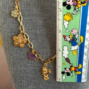 May include: Gold-toned chain necklace with various charms, including a flower, a Mickey Mouse head, a purple bow, and a Mickey Mouse figure. A ruler with Disney characters is in the background. The necklace is displayed on a gray surface.