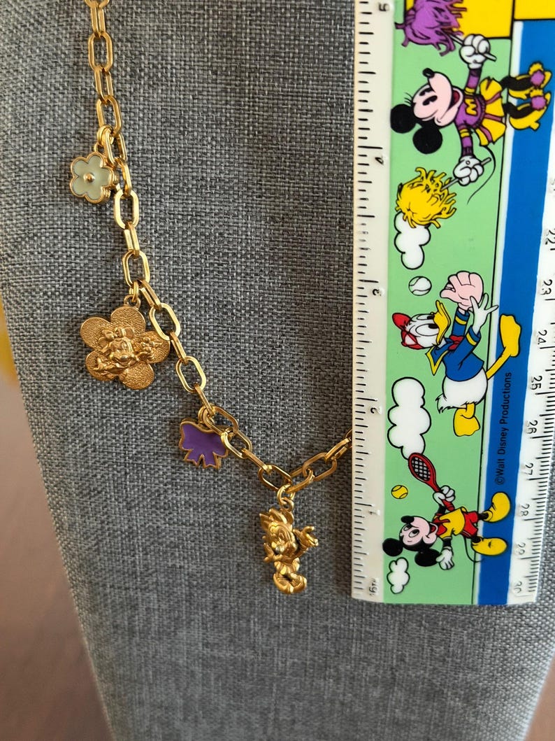 May include: Gold-toned chain necklace with various charms, including a flower, a Mickey Mouse head, a purple bow, and a Mickey Mouse figure. A ruler with Disney characters is in the background. The necklace is displayed on a gray surface.
