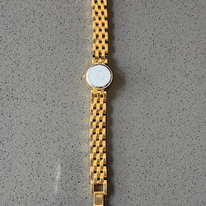 May include: A gold-coloured wristwatch featuring a rectangular link bracelet and a circular watch face. The watch strap is composed of small, interconnected rectangular links. The watch face is white with a silver border. The watch is displayed on a grey surface.
