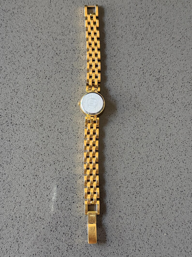 May include: A gold-coloured wristwatch featuring a rectangular link bracelet and a circular watch face. The watch strap is composed of small, interconnected rectangular links. The watch face is white with a silver border. The watch is displayed on a grey surface.
