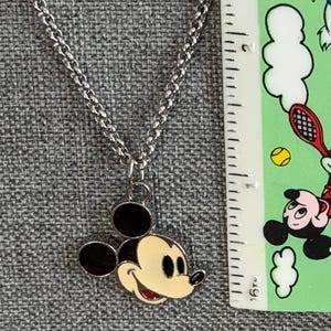 Pu&ograve; includere: Collana color argento con un ciondolo di Topolino. Il ciondolo ha un viso color crema, orecchie nere e bocca rossa. La collana &egrave; mostrata su uno sfondo grigio strutturato.