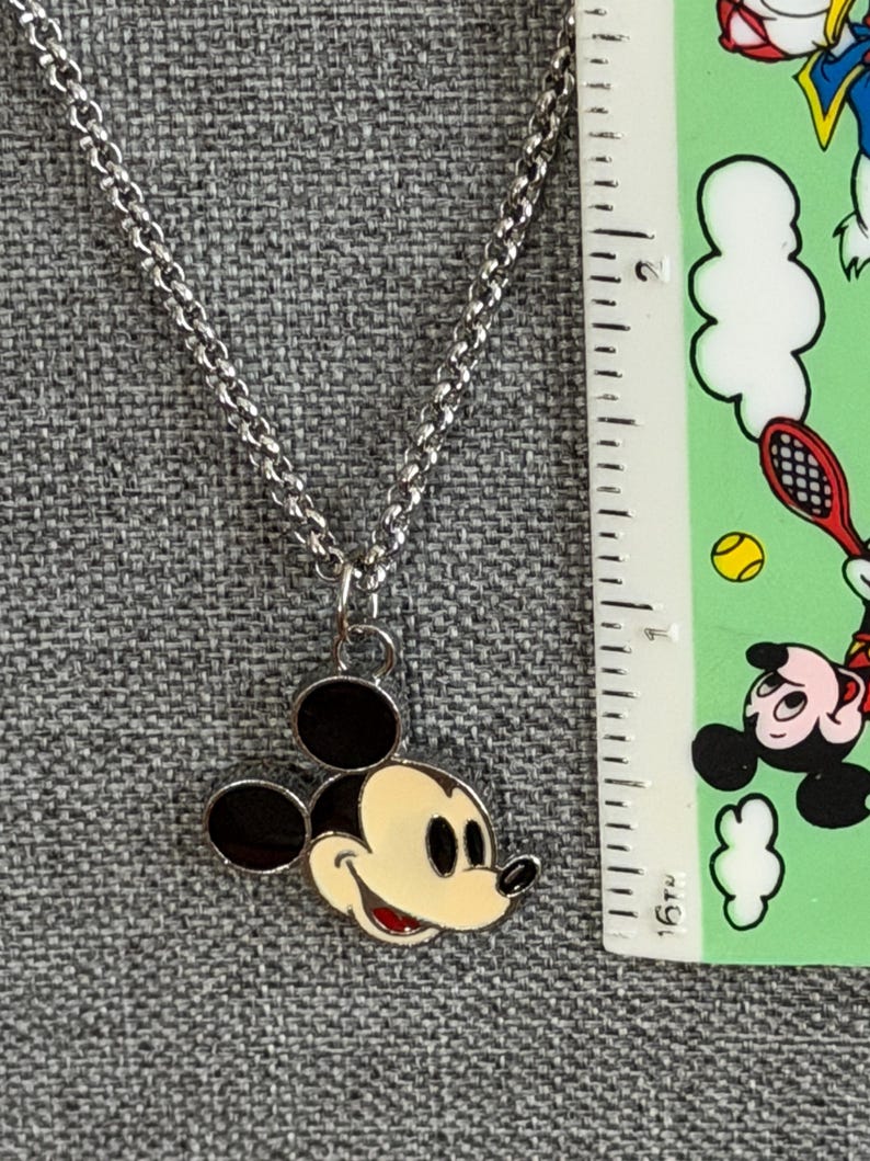Pu&ograve; includere: Collana color argento con un ciondolo di Topolino. Il ciondolo ha un viso color crema, orecchie nere e bocca rossa. La collana &egrave; mostrata su uno sfondo grigio strutturato.