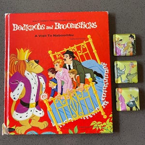 May include: A vintage copy of "Bedknobs and Broomsticks: A Visit To Naboombu" by Walt Disney Productions. The cover is red with illustrations of characters and a bed. Three square magnets with character illustrations are also visible.