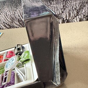 May include: A partially open black watch box with a variety of colorful watch straps and a silver watch with a blue face. The box is on a neutral-colored surface.