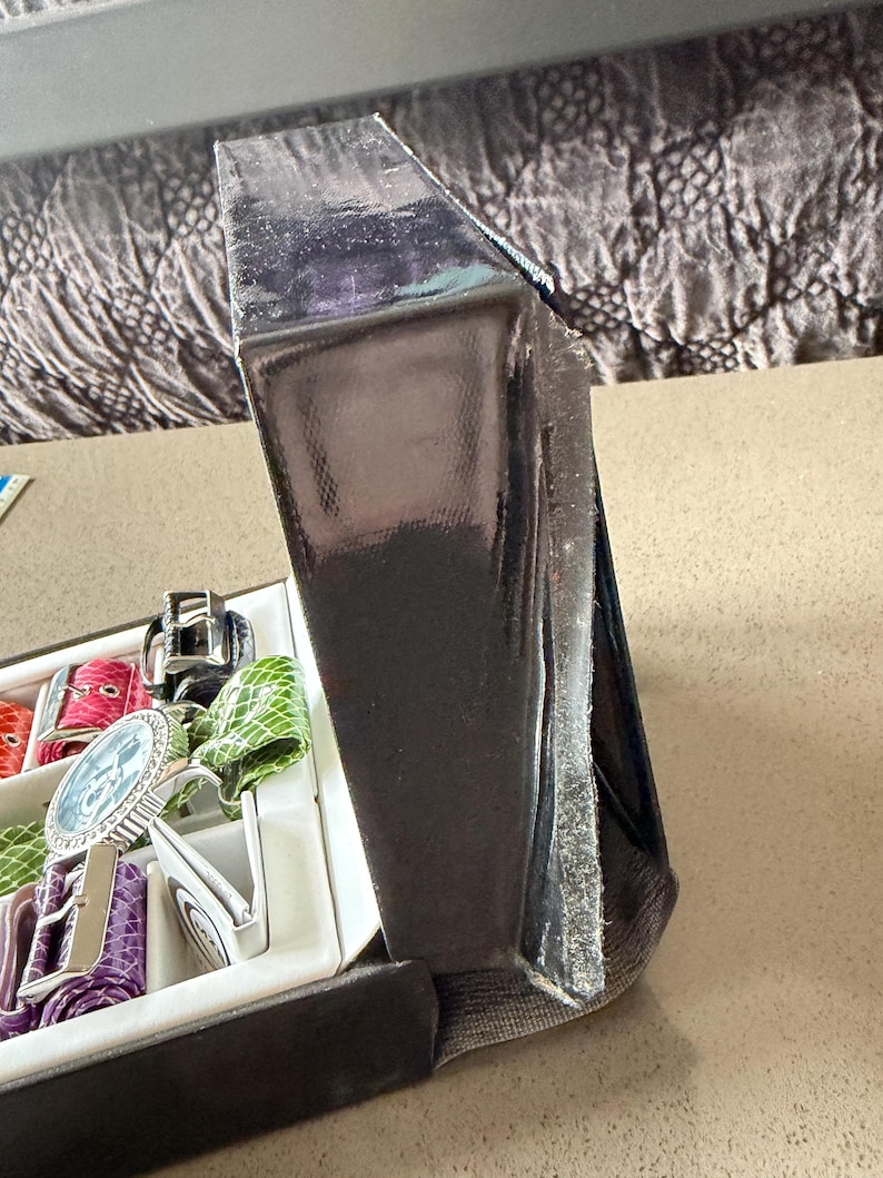 May include: A partially open black watch box with a variety of colorful watch straps and a silver watch with a blue face. The box is on a neutral-colored surface.