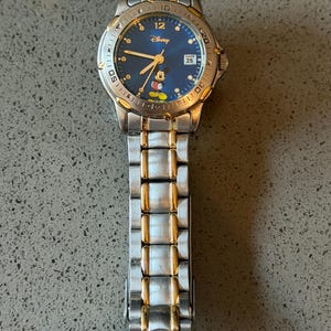 Op de afbeelding: Een zilver- en goudkleurig Disney-horloge met een blauwe wijzerplaat. Het horloge heeft Mickey Mouse, het Disney-logo en een datumweergave. De horlogeband is een combinatie van zilveren en gouden schakels.