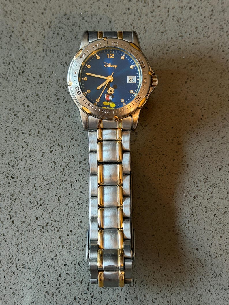 Op de afbeelding: Een zilver- en goudkleurig Disney-horloge met een blauwe wijzerplaat. Het horloge heeft Mickey Mouse, het Disney-logo en een datumweergave. De horlogeband is een combinatie van zilveren en gouden schakels.