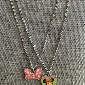 May include: Two silver-toned necklaces. One pendant is a pink bow with white polka dots. The other pendant is a heart-shaped pendant with a Minnie Mouse graphic. The background is a gray textured fabric.