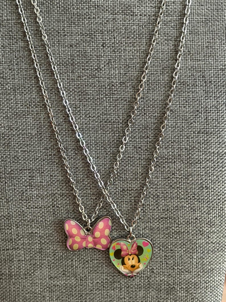 May include: Two silver-toned necklaces. One pendant is a pink bow with white polka dots. The other pendant is a heart-shaped pendant with a Minnie Mouse graphic. The background is a gray textured fabric.