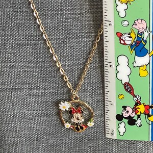 May include: Gold-colored chain necklace with a pendant featuring a cartoon character. The pendant is a circular design with a cartoon character in the center, surrounded by white flowers. The necklace is displayed against a gray background.